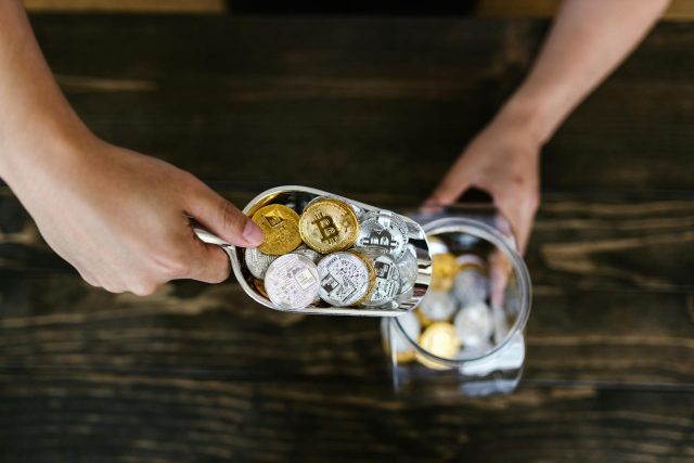 Close-up of hands scooping assorted cryptocurrency coins into a jar on a wooden surface.